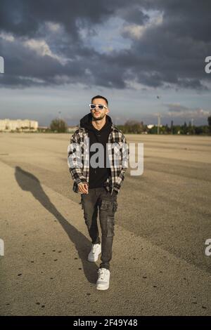 A stylish cool Caucasian male with sunglasses walking on a road in background of gloomy clouds Stock Photo