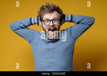 Happy, surprised man screams with joy while holding his hands behind his head. Yellow background. Stock Photo