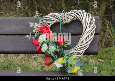 Willow heart partially covered in red roses, foliage and a small wooden butterfly on a wooden bench. Death concept Stock Photo