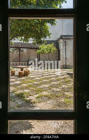 Suzhou, China - May 3, 2010: Closeup through window with black frame of courtyard with weeds and dragon-themed brown decorated stools around circular Stock Photo