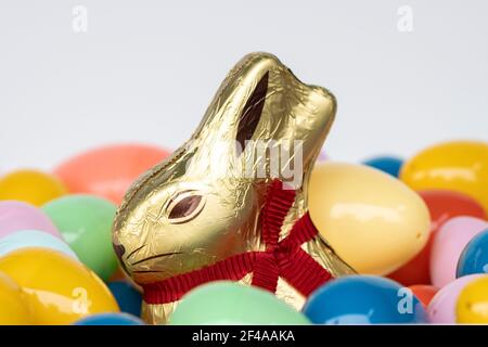 Chocolate Easter bunny wrapped in golden foil with a red ribbon, surrounded by colorful plastic Easter eggs. Stock Photo