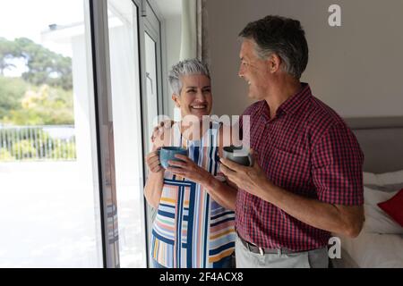 Smiling senior caucasian couple standing by window embracing holding cups of coffee Stock Photo