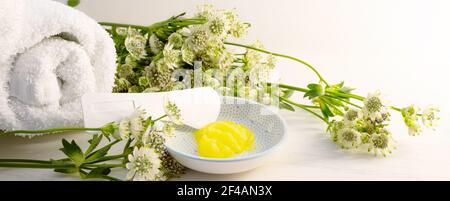 Cosmetic moisturizer or emollient and a spatula for pedicure and soft foot skin in a small bowl and some green flowers on a light wooden background, p Stock Photo