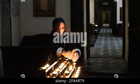 A young Caucasian woman holding her hands over round-shaped tea light burning candles in the Catholic church interior.in Krakow, Poland. Stock Photo