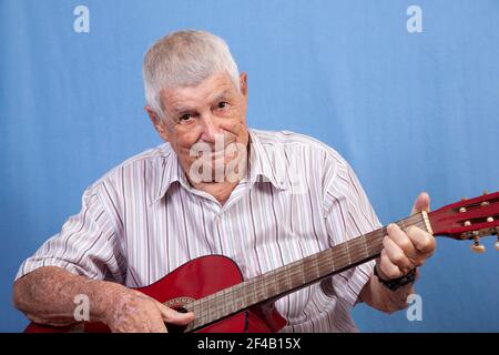 Active happy senior old man with grey hair and a sun damaged skin playing music on a red guitar against a blue background. Stock Photo