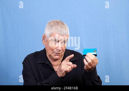 A senior old man with grey hair, sun damaged skin and a hearing aid looking at his credit card or cash card and pointing a finger. Stock Photo