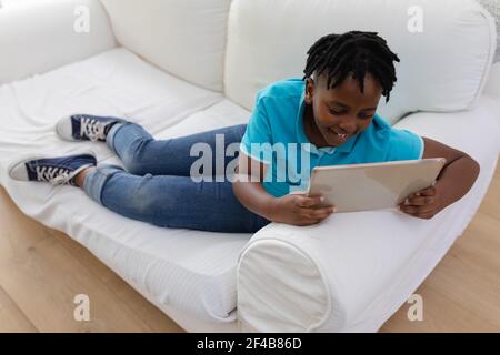 Smiling african american boy with short dreadlocks lying on couch using digital tablet Stock Photo