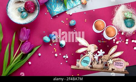 Happy Easter! Painted Easter eggs, decorative wooden bunnies and tulips. Dark/pink background. Top view, panorama banner Stock Photo