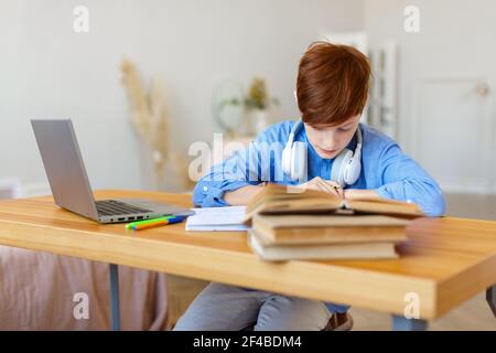 A pupil boy writes an assignment in a notebook during an online lesson in front of a laptop monitor. Online learning concept. Stock Photo