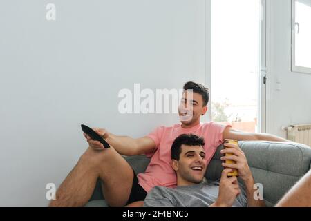 A young Caucasian gay couple relaxing on the couch.  One of them is using his smartphone while the other is using the television remote control Stock Photo