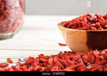 Dried goji berries in wooden bowl, scattered over white boards table under, blurred large glass jar full of fruits in background Stock Photo