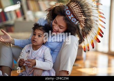 A young mother with indian war bonnet sitting on the floor in a cheerful atmosphere at home and playing with her little daughter. Family, home, playti Stock Photo