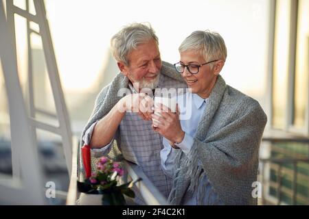 An elderly hugged couple in love warm up with hot tea while enjoying a beautiful sunset from the terrace in a relaxed atmosphere of their apartment. R Stock Photo