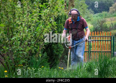 Senior man mowing grass by brushcutter in garden at springtime. Cutting grass in garden. Stock Photo