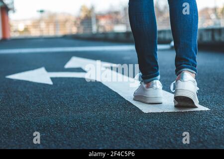 Make decision which way to go. Walking on directional sign on asphalt road. Female legs wearing jeans and white sneakers. Stock Photo