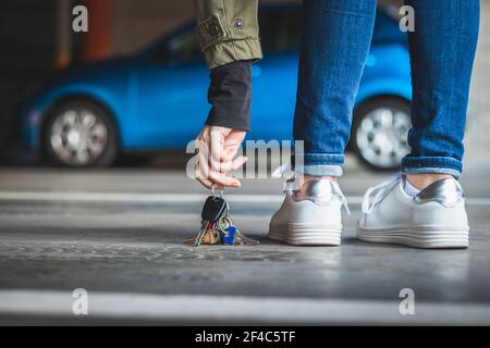 Woman found lost car key at parking lot. Blurry blue car in background. Female legs with white shoes standing at parking garage. Stock Photo