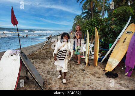 An indigenous Kogi man crosses paths with a surfer on the beach in Palomino, Colombia. Stock Photo