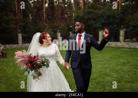 Joyful couple walking at the park, handsome African American man with lovely white woman on wedding day, beautiful bride with charming groom enjoy Stock Photo