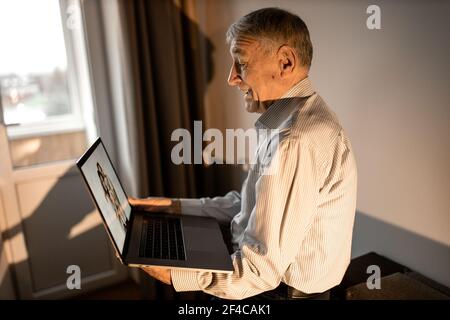 Mature happy man sit on the sofa, looking at the laptop screen. Older male having a video call with grandchildren. Caucasian grandfather use zoom Stock Photo
