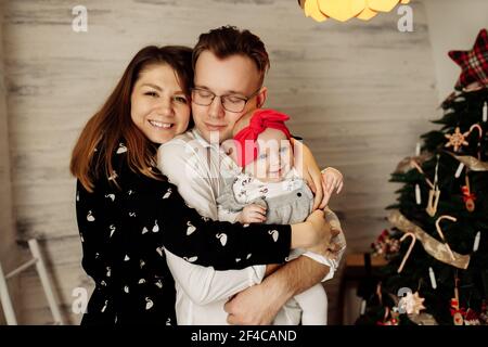 Portrait of happy parents with beautiful baby girl near Christmas tree, handsome father hold little daughter in arms, overjoyed mother smiling, young Stock Photo