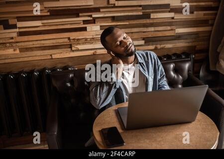 Tired African American businessman taking a break from work. Sits with closed eyes, suffering from neck pain. At the office at the desk with a laptop Stock Photo