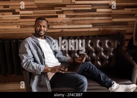 Dreamy smiling African American sitting on sofa with laptop, looking at camera, pondering new opportunities or dreaming of a good future, visualizing Stock Photo