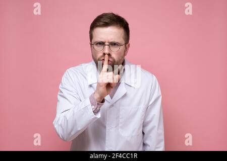 Male doctor with a stethoscope around neck put index finger to lips, making a gesture of silence and looking seriously. Stock Photo
