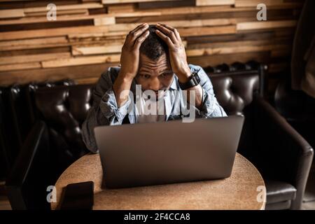 Unhappy young African American man holding his head in hands, tired businessman sitting at work desk with laptop feeling exhausted, financial problems Stock Photo