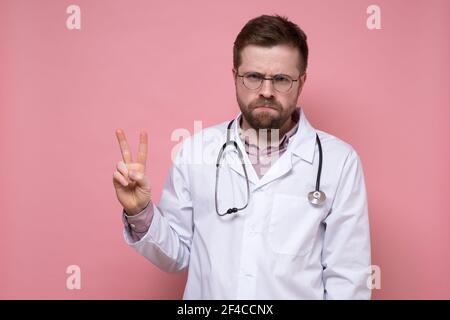 Frustrated male doctor is offended at something, but shows a symbol of victory and frowns. Two fingers gesture.  Stock Photo