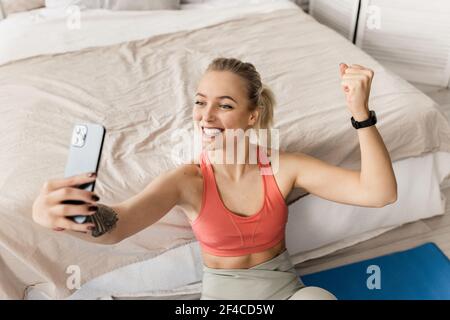 A young yoga instructor sits on a bed and speaks via video communication, resting from workouts, a fit athlete in sportswear uses a mobile phone while Stock Photo