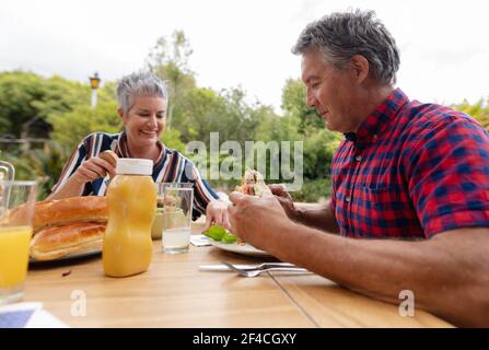 Smiling caucasian senior man holding hamburger eating meal with wife in garden Stock Photo
