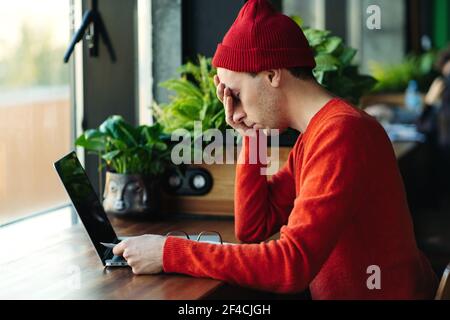 Tired man taking a break during online work on laptop, holding head in hand, feeling lack of energy. Stock Photo