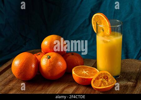 freshly squeezed orange juice in a glass with ice. a pile of oranges and a partially squeezed orange on a wooden board with a teal background. Stock Photo