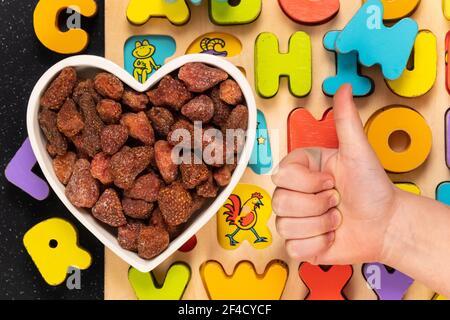 Heap of dried strawberries in a heart shaped bowl with wood block letters. Child hand signaling thumbs up. Healthy snack concept. Stock Photo