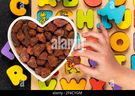 Heap of dried strawberries in a heart shaped bowl with wood block letters. Child hand picking up a strawberry. Healthy snack concept. Stock Photo