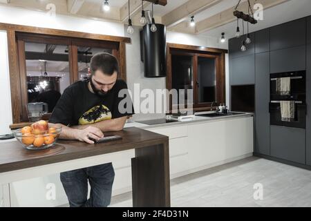 A young Caucasian man standing in the kitchen and playing with his mobile in the window background Stock Photo