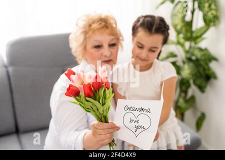 Sincere cute little granddaughter strong cuddling and kissing in cheek her 60s grandmother gave her pretty spring flowers congratulates with birthday Stock Photo