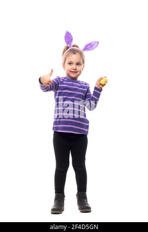 Cute little girl with Easter bunny ears looking up holding painted egg showing thumb up. Full body isolated on white background Stock Photo