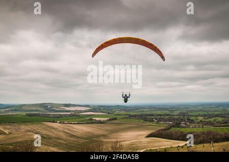 Firle, Lewes, East Sussex, UK. 20th Mar, 2021. Paragliders take to the air under cloudy skies, in the Northerly breeze, from the South Downs with glorious views across the Sussex countryside. Credit: David Burr/Alamy Live News Stock Photo