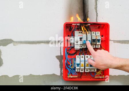 Male hand turns off burning switchboard from overload or short circuit on wall Stock Photo