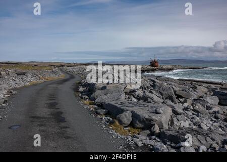 Landscape with grey road, rocky beach, old, rusty shipwreck, blue ocean on a sunny autumn day on Inisheer island, the smallest of Aran islands. Touris Stock Photo