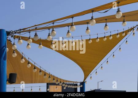 Dynamic texture of fabric stretch roof made of canvas sail with rows of bulb lamps for party night lighting on blue sky background. Stock Photo