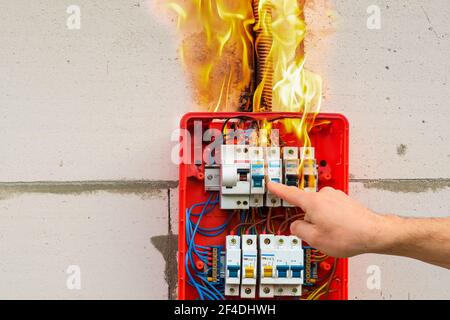 Male hand turns off burning switchboard from overload or short circuit on wall Stock Photo