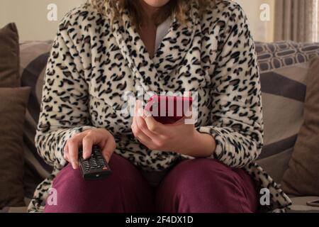 A young lady is sitting on a sofa with a remote tv control and a red smartphone holdings in hands Stock Photo