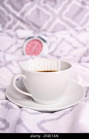 Close up of a coffee cup placed on modern bed sheets with an alarm clock in the background. Morning coffee in bed concept. Morning coffee in bed. Stock Photo