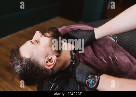 Shaving with straight razor in barbershop, close-up of happy male client leaning back in chair while professional hairdresser does procedure. Stock Photo