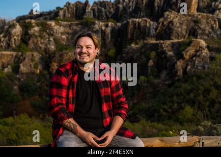 A young Spanish man sitting on a log and smiling on a background of a rocky hill Stock Photo