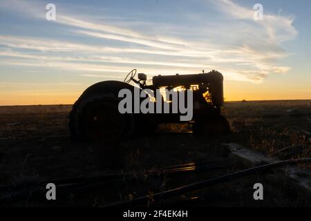 A vintage old farm tractor abandoned in a field silhouetted against a golden sunset in outback Queensland. Stock Photo