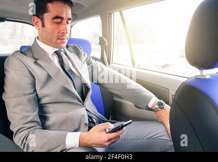 Young handsome businessman sitting in taxi cab while texting sms with smartphone - Business concept with modern man using smart phone Stock Photo