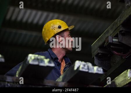 Aluminium electrolysis plant.  Adult Asian worker operator in yellow hardhat and orange earplug on ingot foundry line. Ingots on foreground blurred. Stock Photo